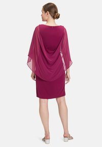 Robe fuchsia avec un superposé transparent drapé à l'arrière. Longueur genoux avec une texture lisse, présentation d'un col rond et d'une fermeture à éclair au dos.