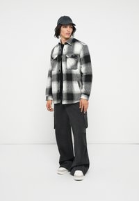 Camicia di flanella a scacchi in bianco e nero, con due tasche sul petto, indossata sopra una maglietta nera; abbinata a pantaloni cargo scuri e sneakers bianche.
