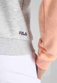 Sudadera a rayas en gris y melocotón con puños acanalados, que presenta un pequeño logo de FILA en rojo, blanco y azul en el dobladillo. Tela suave y texturizada.