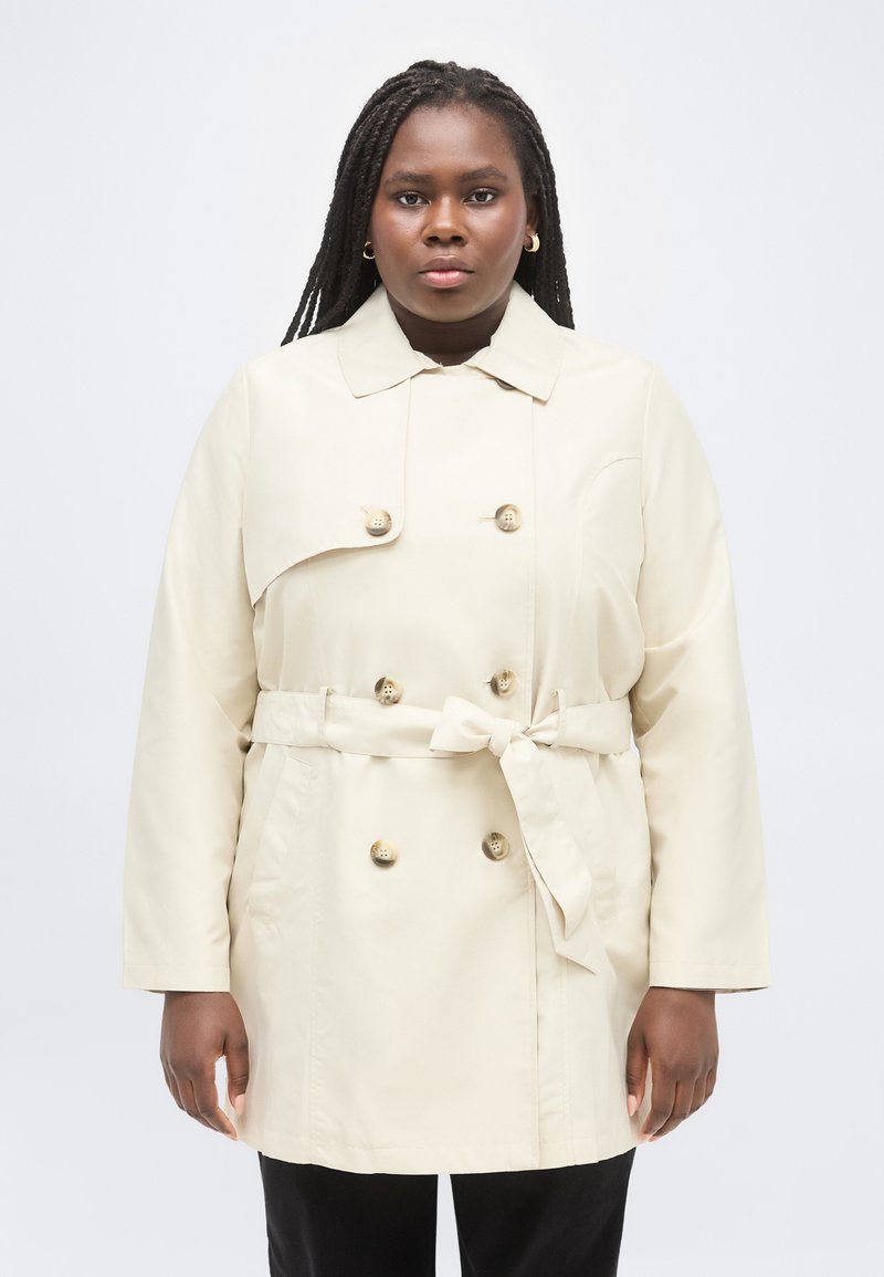 Γυναίκα με μακριά πλεξούδα, που φοράει μια μπεζ διπλόμπουτη καμπαρντίνα με ζώνη δεμένη στη μέση και χρυσά σκουλαρίκια κρίκους, στέκεται μπροστά σε λευκό φόντο.