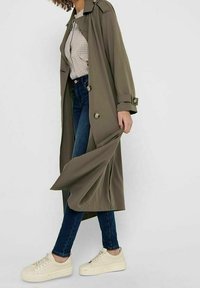 Kobieta w oliwkowym trenczu, w kratkowanej koszuli, ciemnoniebieskich dżinsach i białych sneakersach, trzymająca brzeg płaszcza podczas chodzenia.