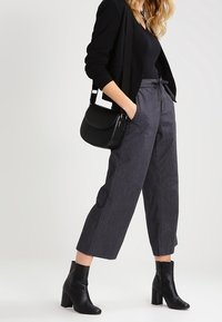 Femme portant un blazer noir, un top noir, un pantalon court gris, des bottines noires, et portant un sac à bandoulière noir, se tenant de profil.