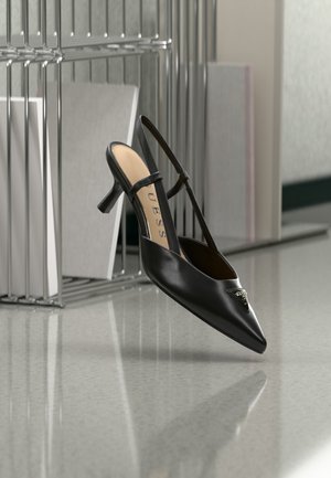 Scarpa nera con tacco kitten slingback, punta appuntita e dettaglio logo in metallo, sospesa vicino a una superficie riflettente e a una rastrelliera metallica con fogli.