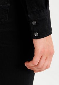Manica di camicia in denim nero con chiusura a bottone in metallo argentato e finitura liscia e strutturata. Il tessuto sembra flessibile e resistente.
