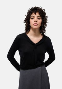 Haut noir à manches longues avec un décolleté drapé et un détail de nœud tordu à l'avant, assorti d'une jupe plissée grise. Texture de tissu lisse.