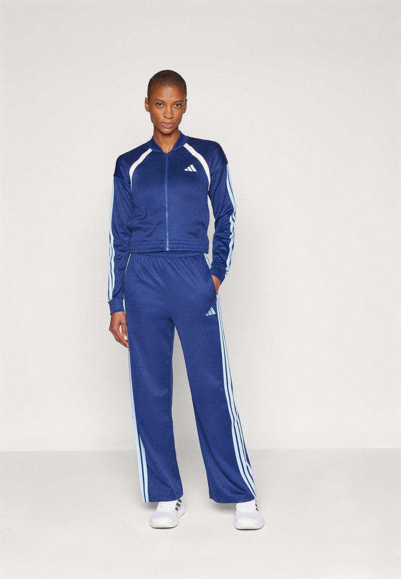 Mørkeblå atletisk treningsdrakt laget av glatt materiale; har hvite tre-stripe detaljer på ermene og buksene, med en zip-up jakke.