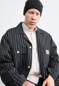 Crna prugasta jakna s kopčanjem na dugmad, dva prednja džepa i Carhartt logo. Nosi se preko svijetle košulje s kragnom i crne kape.