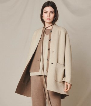 Ung kvinde iført lagdelt beige og brun uldfrakke og cardigan, stående mod en ensfarvet beige baggrund.