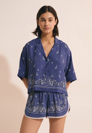 Giovane donna con capelli corti e scuri che indossa una camicia blu ampia a maniche corte con motivo paisley e pantaloncini coordinati con bordo di pizzo.