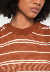 Καφέ ριγέ πλεκτό πουλόβερ με υφασμένη υφή. Διαθέτει λευκές οριζόντιες ρίγες και στρογγυλή λαιμόκοψη, που αναδεικνύουν το στρωματοποιημένο πλεκτό μοτίβο του.