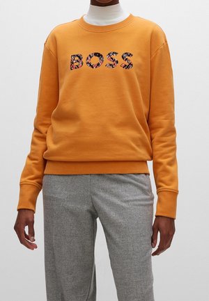 Personne portant un sweat-shirt orange avec le texte floral "BOSS" et un pantalon gris, debout devant un fond clair uni.