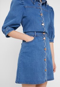 Modrá denimová šaty s krátkými puff rukávy, zapínání na knoflíky vpředu a vroubkovanými detaily podél knoflíků. Obsahuje boční kapsy.