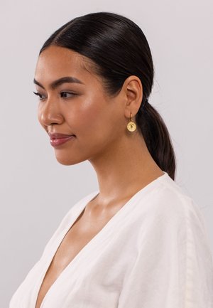 Boucles d'oreilles en or avec un charm circulaire texturé suspendu à chacune. Le modèle porte un vêtement blanc à col en V et a les cheveux raides et foncés.