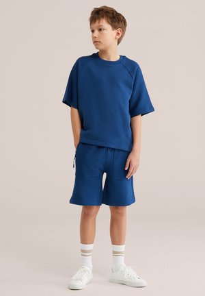 Niño con sudadera de manga corta azul y pantalones cortos a juego, calcetines blancos con rayas beige y zapatillas blancas, de pie con las manos relajadas.