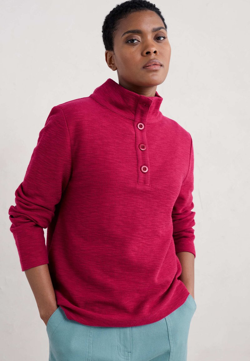 Pullover fucsia con colletto alto, placket a tre bottoni e maniche lunghe, realizzato in tessuto testurizzato, abbinato a pantaloni azzurri chiari.