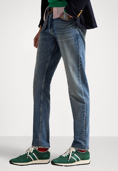 Jeans in denim blu con gamba dritta e texture sbiadita, indossati con sneakers verdi con strisce bianche, e sovrapposti a un top verde e rosa.