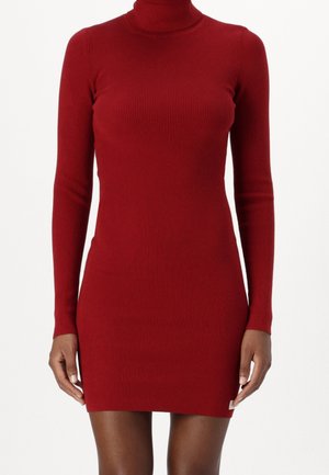 Vartaloa myötäilevä punainen, ribbineulottu poolomekko, jossa pitkä hiha, päällä, ylettyen reiden keskivaiheille, pienellä valkoisella lipulla helmassa.