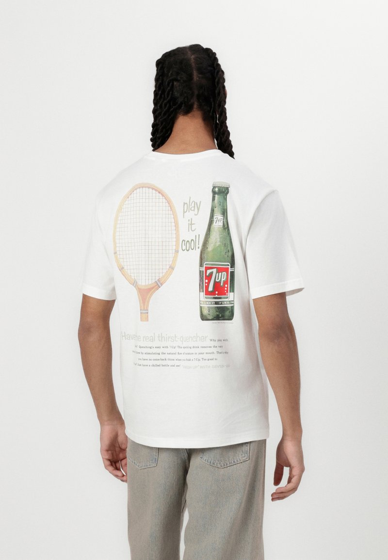 Hvid bomulds t-shirt med et grafisk design på ryggen, der viser et tennisracket og en flaske 7-Up. Teksten inkluderer "spil det cool!" og et slogan.