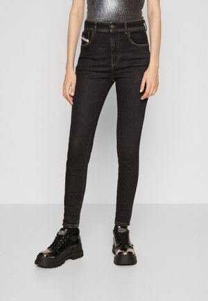 Person iført sorte skinny jeans og sorte platformsneakers med metalspidser, stående mod en ensfarvet grå baggrund.