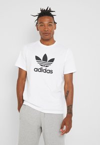 Fehér pamut póló fekete Adidas logóval, három csíkkal; kerek nyakú kialakítás és rövid ujjú. Szürke meLegítőnadrággal párosítva.
