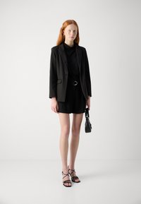 Black blazer over a black top, paired with a black belt and shorts. Black strappy sandals and a small black handbag. Smooth textures.