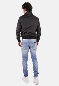 Giacca bomber nera con colletto alto, polsini a coste, abbinata a jeans slim-fit blu chiaro e sneakers bianche, vista da dietro.