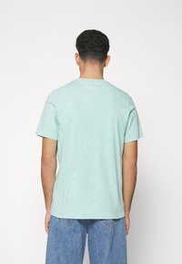 Mentově zelené bavlněné tričko, kulatý výstřih, krátké rukávy, lehká textura, s jemnými variacemi v barvě, nošeno s modrými džíny.