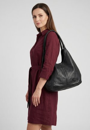 Frau in einem burgunderroten Kleid, die eine große schwarze Lederschultertasche vor einem schlichten hellen Hintergrund trägt.