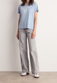 Lichtblauwe katoenen t-shirt met korte mouwen en een logo, gecombineerd met hoog opgesneden, wijde grijze jeans en witte sneakers.