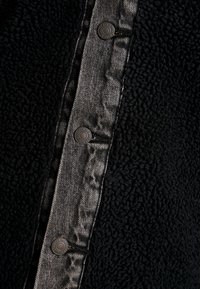 Musta fleece-takki, jossa on farkkutasku kiiltävillä nappeilla, tekstuurinen materiaali, kontrastinen kiilto ja monimutkainen nappimuotoilu.