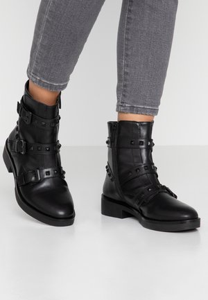 Osoba nosząca czarne skórzane botki z klamrami i ćwiekami, w połączeniu z dopasowanymi szarymi dżinsami na jednolitym tle.