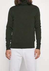 Ciemnozielony sweter w serek wykonany z miękkiego materiału dzianinowego. Dopasowany krój z długimi rękawami i prążkowanymi mankietami. Nosi się z białymi spodniami.