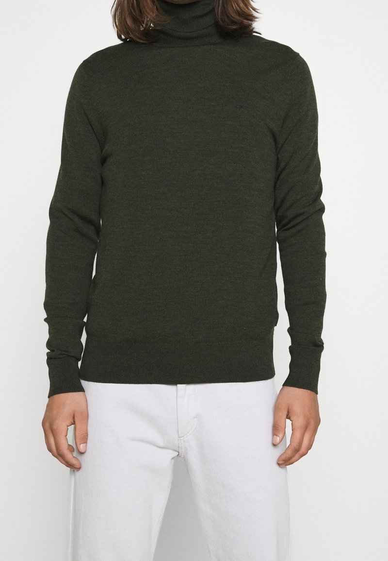 Ciemnozielony sweter w serek wykonany z miękkiego materiału dzianinowego. Dopasowany krój z długimi rękawami i prążkowanymi mankietami. Nosi się z białymi spodniami.