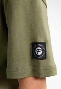 Camiseta de algodón verde oliva con una etiqueta negra en la manga que presenta un logotipo circular y texto. Textura suave y mangas cortas.