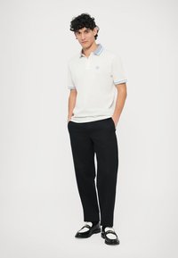 Hvit poloshirt med lyseblå krage og ribbestrikkede mansjetter, kombinert med svarte bukser og blanke loafers med hvite detaljer.