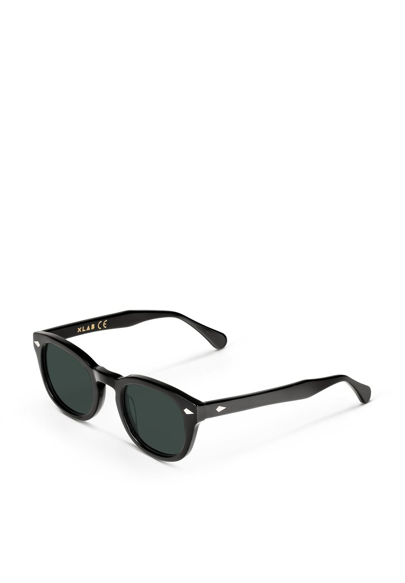 Gafas de sol negras con un marco grueso. Lentes oscuros en forma ovalada y acentos en forma de diamante en las varillas. Acabado texturizado en el marco.