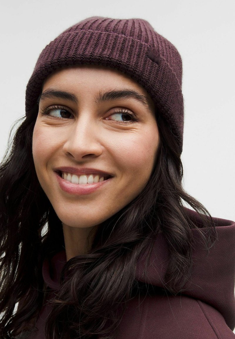 Joven mujer con cabello oscuro sonriendo, vistiendo un gorro de punto acanalado color ciruela y una sudadera a juego, mirando hacia la izquierda sobre un fondo liso.