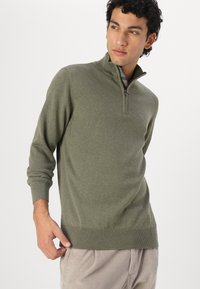 Olivgrüner Pullover mit Reißverschluss am Hals, aus Strickstoff, mit gerippten Bündchen und Saum sowie einer strukturierten Oberfläche. Mit einem dezenten Logo auf der Brust.