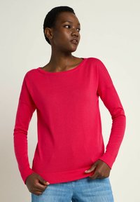 Pink, long-sleeve sweater with a wide neckline, crafted from soft fabric. Features a loose fit and ribbed hem, complemented by denim bottoms.