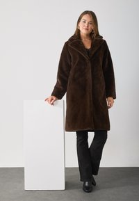 Brown faux fur overcoat with a wide collar, long sleeves, and a straight cut. Worn over black pants and ankle boots against a white backdrop.