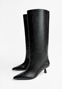 Bottes en cuir noir arrivee genoux avec un bout pointu et un petit talon bloc. Elles présentent une texture lisse et un design élégant.