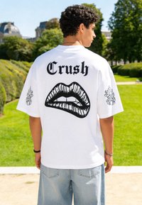 Jeune homme en t-shirt blanc avec le texte « Crush » et un graphisme noir de lèvres dans le dos, debout à l'extérieur dans un parc verdoyant.