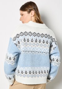 Jasnoniebieski sweter z białym kołnierzykiem i misternym wzorem dzianiny. Ma zdobne wzory w czerni i szarości wokół ramion.