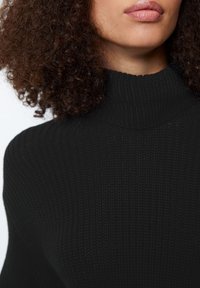 Maglione a collo alto nero a coste, con una scollatura alta e tessuto testurizzato. Vista ravvicinata che enfatizza il materiale e la struttura.