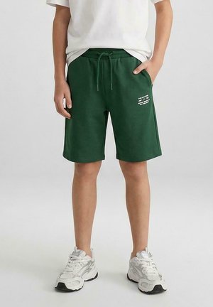 Kind draagt donker groene korte broek met trekkoord en witte tekst op linkerpijp, wit t-shirt en witte sneakers, staat met handen deels in de zakken.
