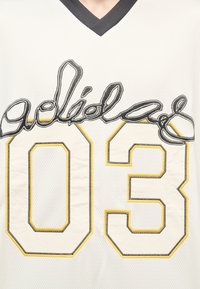 Λευκό αθλητικό πουκάμισο με μαύρες και κίτρινες λεπτομέρειες, με κεντημένο κείμενο και μεγάλο ραμμένο αριθμό 03 στο μπροστινό μέρος.