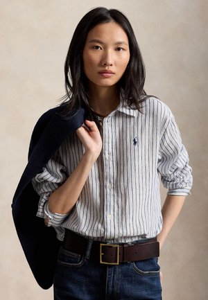 Femme aux longs cheveux foncés porte une chemise à rayures bleues et blanches boutonnée, un jean foncé et tient une veste sombre sur son épaule.