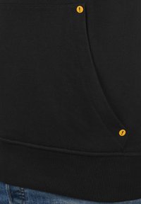 Zwarte sweater met een zachte textuur, voorzien van een zak met gele accentknopen en een geribd zoom. Jeans zijn zichtbaar aan de onderkant.