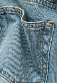 Gros plan sur un tissu denim bleu clair montrant une poche pliée avec des coutures orange visibles et un motif de tissage texturé.