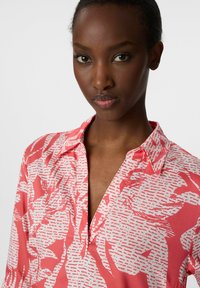 Różowa bluzka z kołnierzykiem, z białym abstrakcyjnym wzorem. Miękka tkanina z luźnym krojem i półzapięciem przy dekolcie.